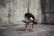 A person is performing a yoga pose, balancing on their hands with knees tucked close to the arms, against a textured stone wall. The individual has gray hair tied back and is wearing a black top and gray leggings. The setting appears to be outdoors on a brick platform.