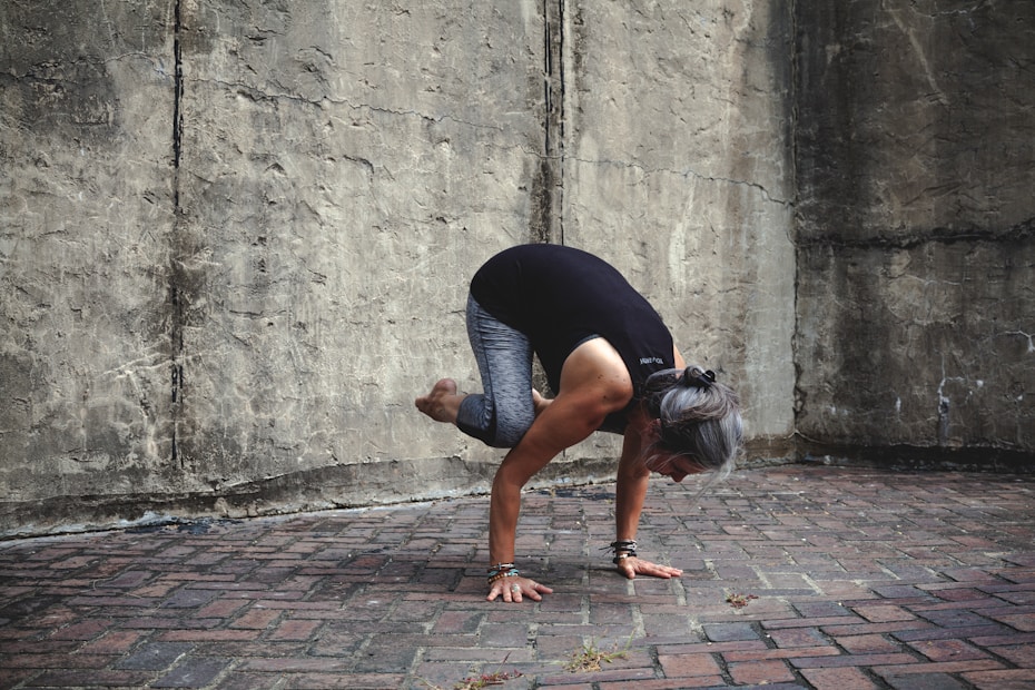 A person is performing a yoga pose, balancing on their hands with knees tucked close to the arms, against a textured stone wall. The individual has gray hair tied back and is wearing a black top and gray leggings. The setting appears to be outdoors on a brick platform.