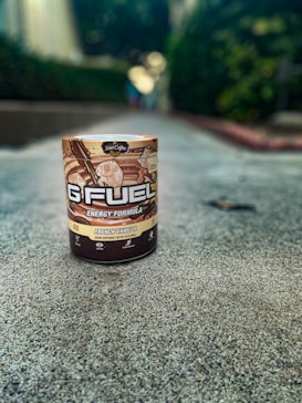 A container of G Fuel energy formula with French vanilla flavor placed on a textured stone or pavement surface. The background is blurred but shows green foliage and outdoor scenery.