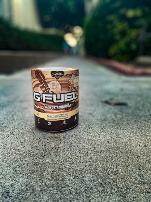 A container of G Fuel energy formula with French vanilla flavor placed on a textured stone or pavement surface. The background is blurred but shows green foliage and outdoor scenery.