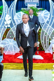 A man in a formal black suit with a white shirt walks down the aisle of a venue decorated with white floral arrangements and a red carpet. Behind him, a man stands at a podium with the phrase 'DEUS É AMOR' displayed, surrounded by white netted decorations and green plants.