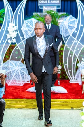 A man in a formal black suit with a white shirt walks down the aisle of a venue decorated with white floral arrangements and a red carpet. Behind him, a man stands at a podium with the phrase 'DEUS É AMOR' displayed, surrounded by white netted decorations and green plants.
