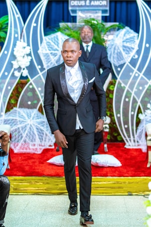 A man in a formal black suit with a white shirt walks down the aisle of a venue decorated with white floral arrangements and a red carpet. Behind him, a man stands at a podium with the phrase 'DEUS &Eacute; AMOR' displayed, surrounded by white netted decorations and green plants.