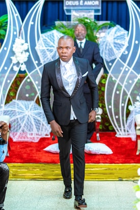 A man in a formal black suit with a white shirt walks down the aisle of a venue decorated with white floral arrangements and a red carpet. Behind him, a man stands at a podium with the phrase 'DEUS &Eacute; AMOR' displayed, surrounded by white netted decorations and green plants.
