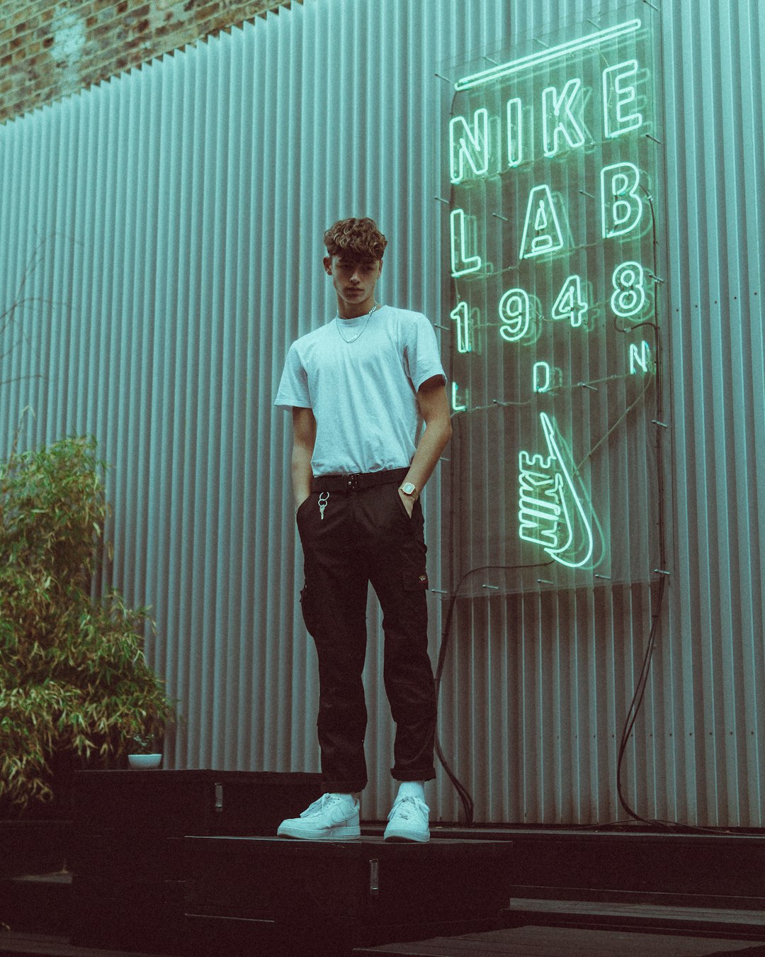 man in white t-shirt and black pants standing near green wall during daytime