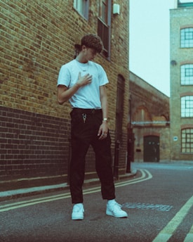 A young person stands in an urban alley, wearing a white t-shirt, dark pants, and white sneakers. The surrounding environment features brick buildings and a cobblestone street.