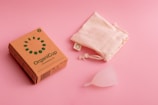 A cardboard box labeled 'OrganiCup' with green lettering lies on a pink surface next to a drawstring cloth bag and a translucent menstrual cup. The box has additional small icons and text, indicating product details.