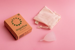 A cardboard box labeled 'OrganiCup' with green lettering lies on a pink surface next to a drawstring cloth bag and a translucent menstrual cup. The box has additional small icons and text, indicating product details.