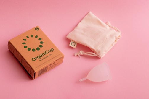 A cardboard box labeled 'OrganiCup' with green lettering lies on a pink surface next to a drawstring cloth bag and a translucent menstrual cup. The box has additional small icons and text, indicating product details.