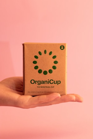 A hand is holding a brown cardboard box against a pink background. The box features green text and a circular pattern, with the brand name 'OrganiCup' and the description 'The Menstrual Cup'.