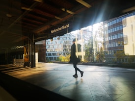 A person is walking through a train station platform, with the sign 'Hackescher Markt' visible above. The sunlight creates reflections and shadows on the shiny floor, and the background showcases modern buildings with large glass windows.