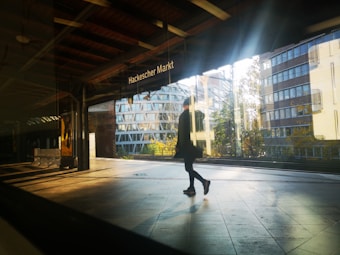 A person is walking through a train station platform, with the sign 'Hackescher Markt' visible above. The sunlight creates reflections and shadows on the shiny floor, and the background showcases modern buildings with large glass windows.