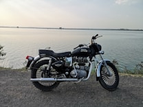 A black Royal Enfield motorcycle parked by a serene lakeside. The horizon stretches out with a calm body of water under a slightly overcast sky. Sparse vegetation can be seen along the shoreline, with distant trees and landmass barely visible.