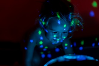 A child surrounded by colorful projections of glowing stars and dots, with an expression of wonder. The setting is dark, highlighting the vibrant hues of green, blue, and yellow.