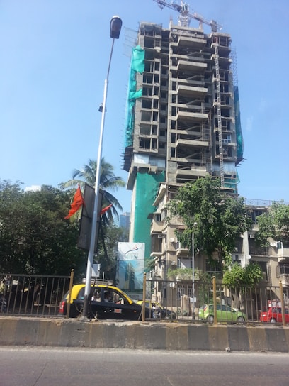 A tall building under construction is surrounded by scaffolding and green netting. In the foreground, a black and yellow taxi is parked beside a billboard. Palm trees and other greenery are visible near the base of the building. A clear blue sky dominates the background, and a streetlamp is visible on the left side of the image.