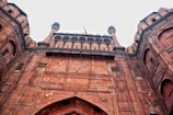 A large, imposing fort with detailed architecture, characterized by massive red sandstone walls and two grand towers. Small intricate windows and decorative carvings are visible. An Indian flag is flying atop the fort.
