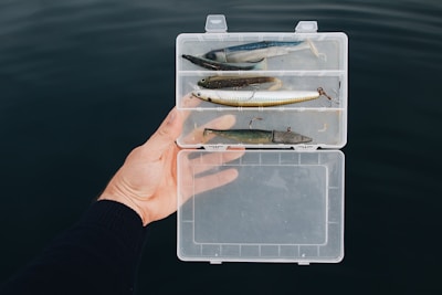 A person holds a transparent plastic tackle box containing several fishing lures over a dark water background. The lures vary in color and size, resting in separate compartments, and the person's hand is visible wearing a dark sleeve.