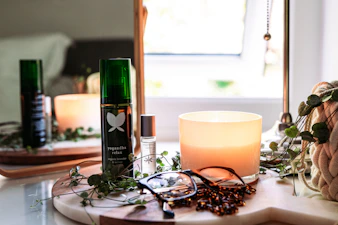 A cozy setup featuring a lit candle, a green bottle labeled 'yogandha relax', a smaller bottle with a rollerball top, a pair of eyeglasses, and a plant in the background. The items are placed on a wooden tray with a soft curtain of natural light entering through a nearby window, contributing to a serene and relaxing atmosphere.