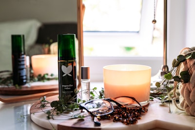 A cozy setup featuring a lit candle, a green bottle labeled 'yogandha relax', a smaller bottle with a rollerball top, a pair of eyeglasses, and a plant in the background. The items are placed on a wooden tray with a soft curtain of natural light entering through a nearby window, contributing to a serene and relaxing atmosphere.