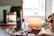 A cozy setup featuring a lit candle, a green bottle labeled 'yogandha relax', a smaller bottle with a rollerball top, a pair of eyeglasses, and a plant in the background. The items are placed on a wooden tray with a soft curtain of natural light entering through a nearby window, contributing to a serene and relaxing atmosphere.