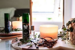 A cozy setup featuring a lit candle, a green bottle labeled 'yogandha relax', a smaller bottle with a rollerball top, a pair of eyeglasses, and a plant in the background. The items are placed on a wooden tray with a soft curtain of natural light entering through a nearby window, contributing to a serene and relaxing atmosphere.