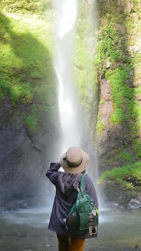 A person wearing a wide-brimmed hat and a green backpack stands facing a tall waterfall surrounded by lush greenery. The water cascades down a rock face into a pool, creating a serene and refreshing atmosphere.