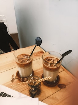 Two iced coffee drinks served in clear glasses with black spoons resting inside. The drinks are placed on a light wooden table along with a small glass vase containing dried flowers. Part of a laptop is visible in the bottom corner, suggesting a casual setting.