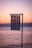 A Greek flag is gently waving in the foreground against a serene backdrop of a calm sea and a pastel-colored sunset sky. The horizon shows a faint outline of distant mountains bathed in soft evening light.