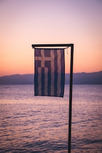 A Greek flag is gently waving in the foreground against a serene backdrop of a calm sea and a pastel-colored sunset sky. The horizon shows a faint outline of distant mountains bathed in soft evening light.