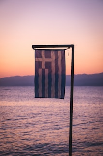 A Greek flag is gently waving in the foreground against a serene backdrop of a calm sea and a pastel-colored sunset sky. The horizon shows a faint outline of distant mountains bathed in soft evening light.