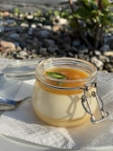 A jar of creamy dessert topped with a layer of orange fruit sauce and a mint leaf, placed on a white paper napkin with a metal spoon beside it. The background consists of pebbles and some greenery, indicating an outdoor setting.