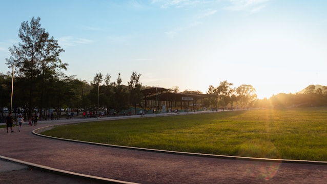 A park setting with a paved path curving through a grassy field. Several trees line the path, and people are walking and gathering. A sunlit sky enhances the scene with a warm glow.