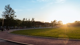 A park setting with a paved path curving through a grassy field. Several trees line the path, and people are walking and gathering. A sunlit sky enhances the scene with a warm glow.