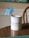 A white mug with a green and blue logo and the words 'GoWP Create Happiness' printed on it sits on a wooden table. A blue cocktail umbrella and a pink straw are placed inside the mug.