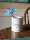 A white mug with a green and blue logo and the words 'GoWP Create Happiness' printed on it sits on a wooden table. A blue cocktail umbrella and a pink straw are placed inside the mug.
