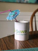 A white mug with a green and blue logo and the words 'GoWP Create Happiness' printed on it sits on a wooden table. A blue cocktail umbrella and a pink straw are placed inside the mug.