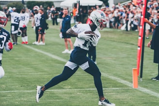 A football player wearing a helmet and uniform is running on a grass field while holding a football tightly. Surrounding him are other players and coaches, with a crowd of spectators in the background. The scene appears to be a practice or training session with people observing the action.