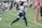 A football player wearing a helmet and uniform is running on a grass field while holding a football tightly. Surrounding him are other players and coaches, with a crowd of spectators in the background. The scene appears to be a practice or training session with people observing the action.