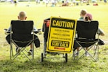 A group of people sitting in folding chairs on a grassy field facing a sports event. A prominent yellow sign reads, 'Caution: Players Playing, Coaches Coaching, Officials Officiating, Mistakes Will Be Made, Stay Calm'. The sign is attached to a cart and positioned between the chairs.