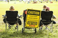 A group of people sitting in folding chairs on a grassy field facing a sports event. A prominent yellow sign reads, 'Caution: Players Playing, Coaches Coaching, Officials Officiating, Mistakes Will Be Made, Stay Calm'. The sign is attached to a cart and positioned between the chairs.