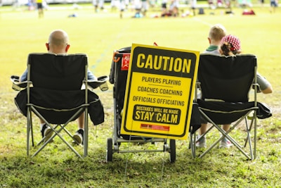 A group of people sitting in folding chairs on a grassy field facing a sports event. A prominent yellow sign reads, 'Caution: Players Playing, Coaches Coaching, Officials Officiating, Mistakes Will Be Made, Stay Calm'. The sign is attached to a cart and positioned between the chairs.