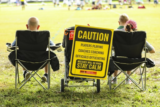 A group of people sitting in folding chairs on a grassy field facing a sports event. A prominent yellow sign reads, 'Caution: Players Playing, Coaches Coaching, Officials Officiating, Mistakes Will Be Made, Stay Calm'. The sign is attached to a cart and positioned between the chairs.