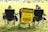 A group of people sitting in folding chairs on a grassy field facing a sports event. A prominent yellow sign reads, 'Caution: Players Playing, Coaches Coaching, Officials Officiating, Mistakes Will Be Made, Stay Calm'. The sign is attached to a cart and positioned between the chairs.
