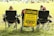 A group of people sitting in folding chairs on a grassy field facing a sports event. A prominent yellow sign reads, 'Caution: Players Playing, Coaches Coaching, Officials Officiating, Mistakes Will Be Made, Stay Calm'. The sign is attached to a cart and positioned between the chairs.