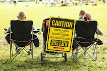 A group of people sitting in folding chairs on a grassy field facing a sports event. A prominent yellow sign reads, 'Caution: Players Playing, Coaches Coaching, Officials Officiating, Mistakes Will Be Made, Stay Calm'. The sign is attached to a cart and positioned between the chairs.