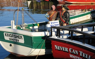 A small boat labeled 'KINGFISHER WHITBY' is docked in a marina. Two pirate mannequins dressed in traditional pirate attire stand on the boat, with a sign promoting 'Kingfisher Sea Trips'. The water reflects the surrounding boats and harbor.