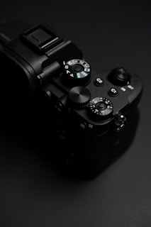A close-up view of a black digital camera with visible dials and controls on a dark background, showcasing its intricate features and ergonomic design.