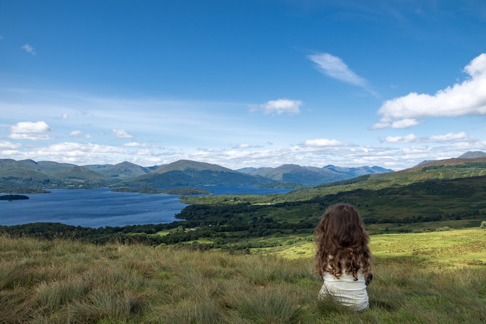 woman in white shirt sitting on green grass field near body of water during daytime