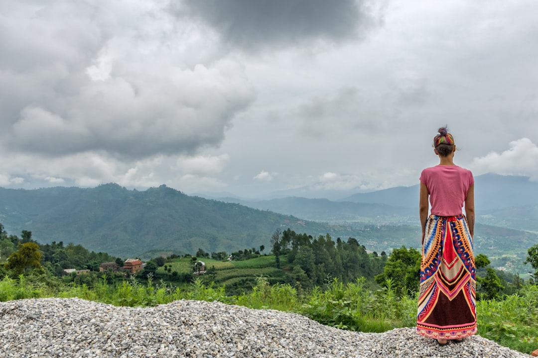 man in red tank top standing on rocky hill looking at green mountains under white cloudy,