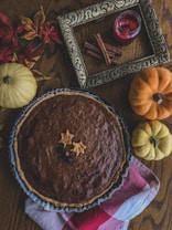 A rustic autumn display features a pumpkin pie on a wooden table, surrounded by small yellow and orange pumpkins, colorful fall leaves, a decorative frame, a red candle, and cinnamon sticks. A red and white checkered cloth is partially visible under the pie.